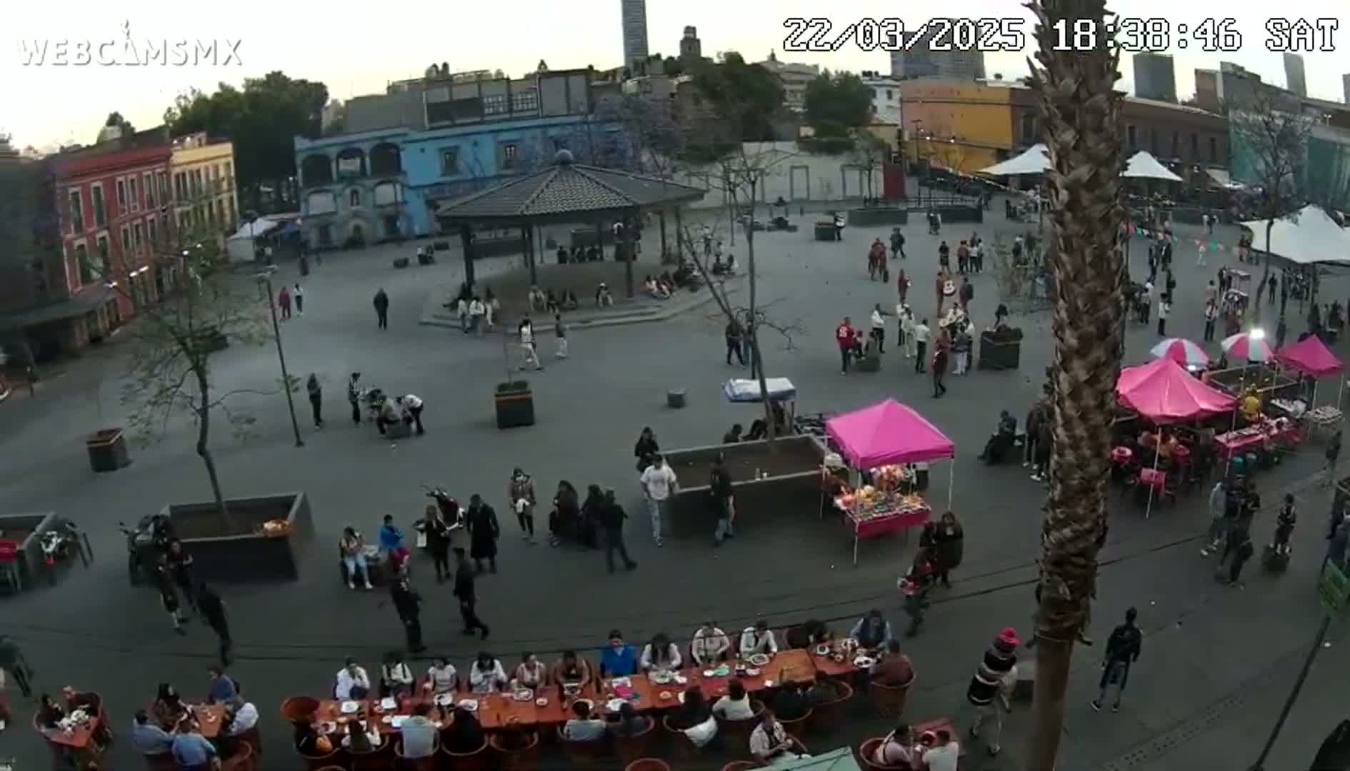 A large plaza with numerous people gathered around food stalls and tables under a mostly clear evening sky.