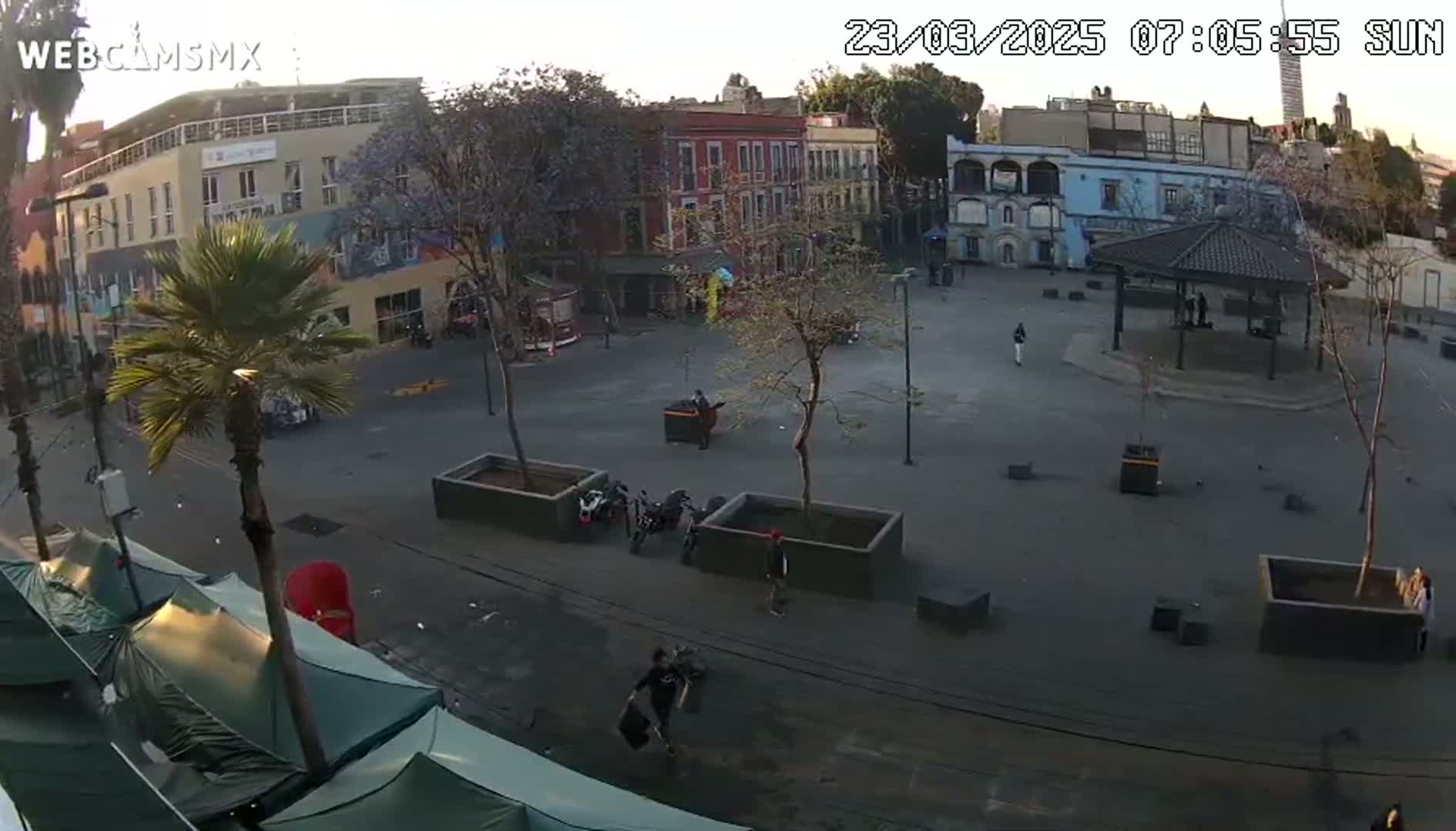 A mostly empty plaza with several buildings, trees, and a gazebo is seen on a partly sunny morning.