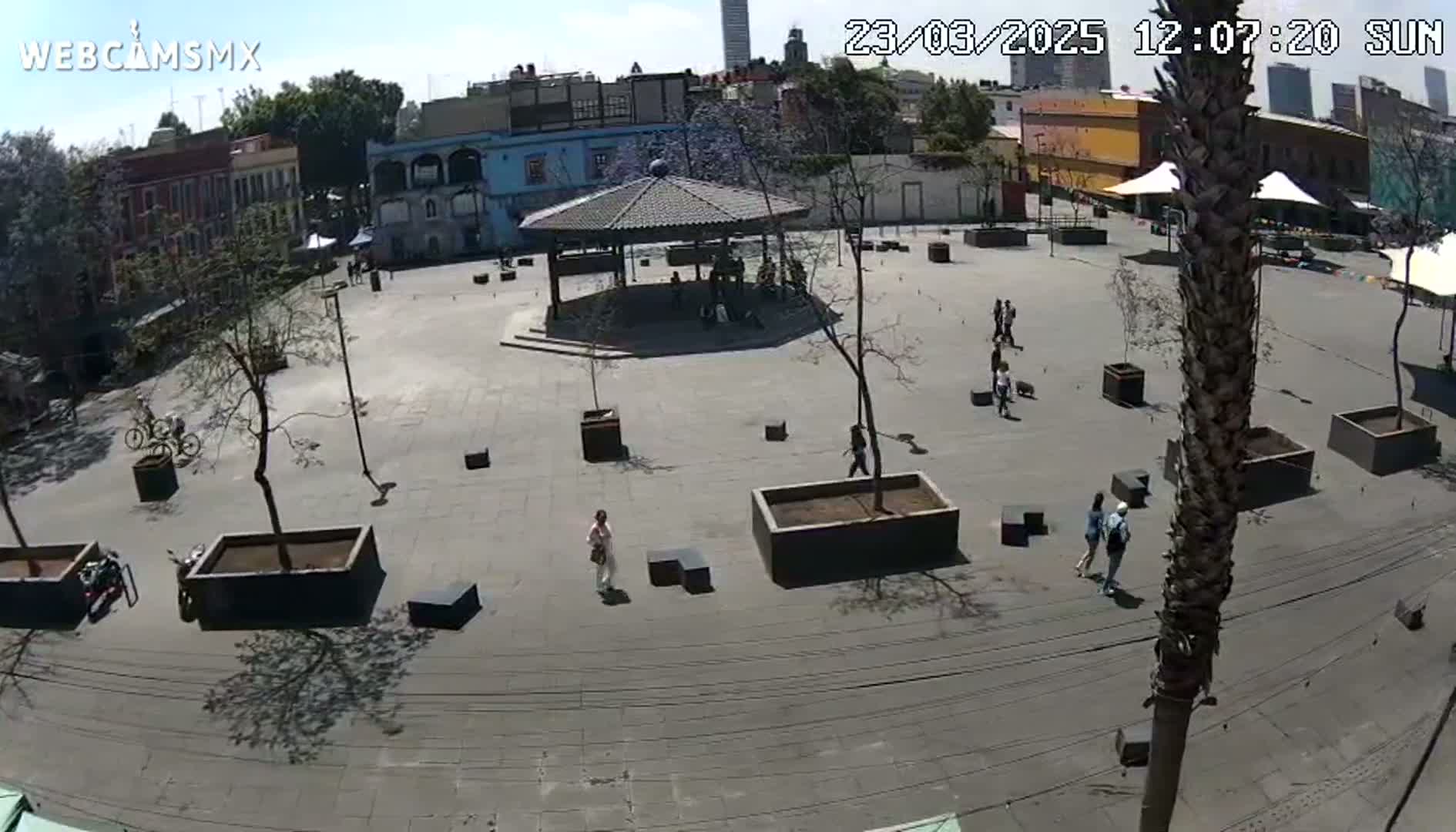 A sunny outdoor plaza with a gazebo, several people walking, and numerous potted plants.