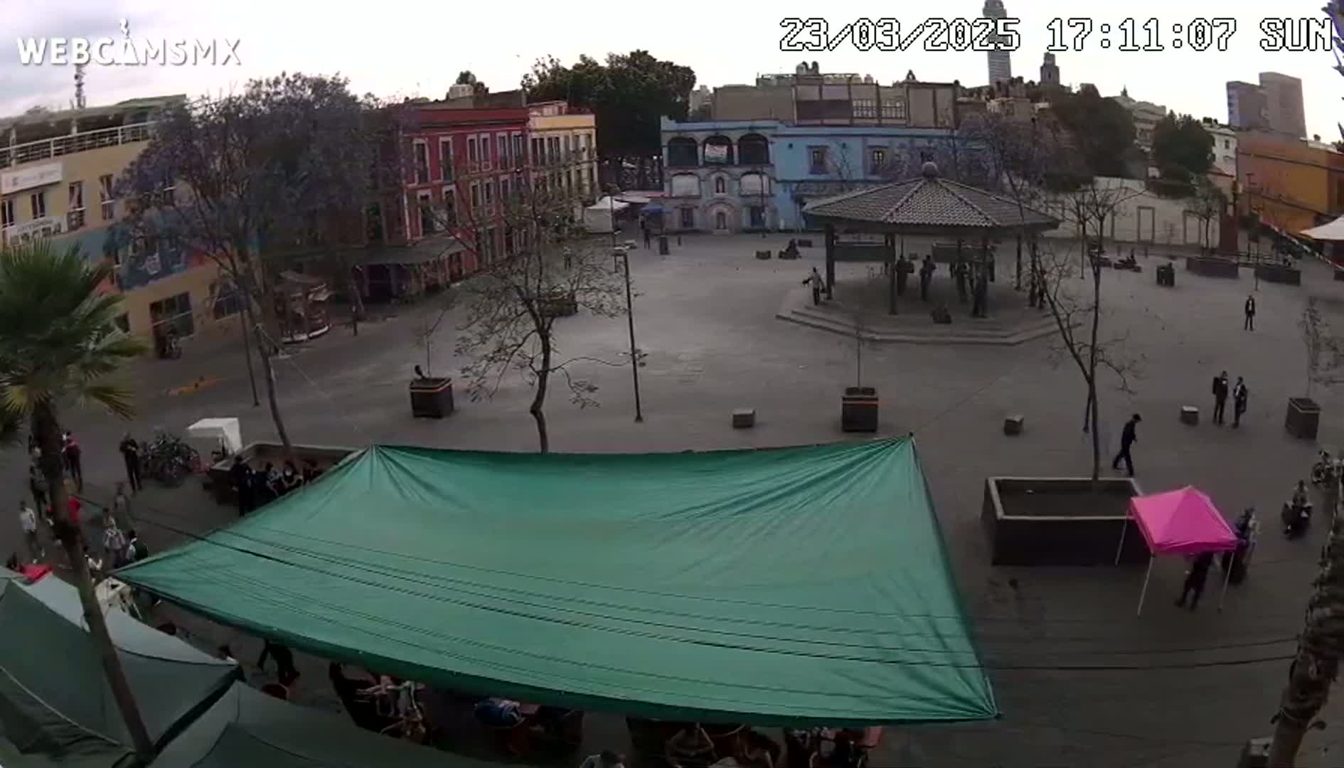 A mostly empty town square with a gazebo, several buildings, and a green canopy, under an overcast sky.