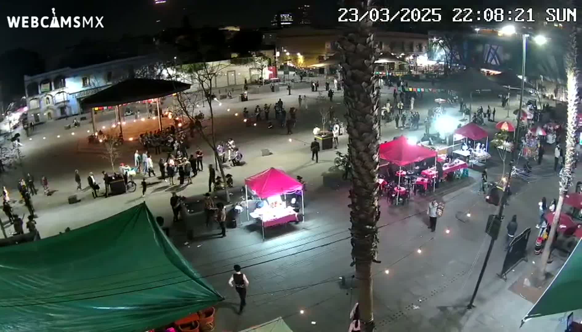 A nighttime, outdoor plaza scene shows numerous people walking among market stalls and small tents under bright lights.