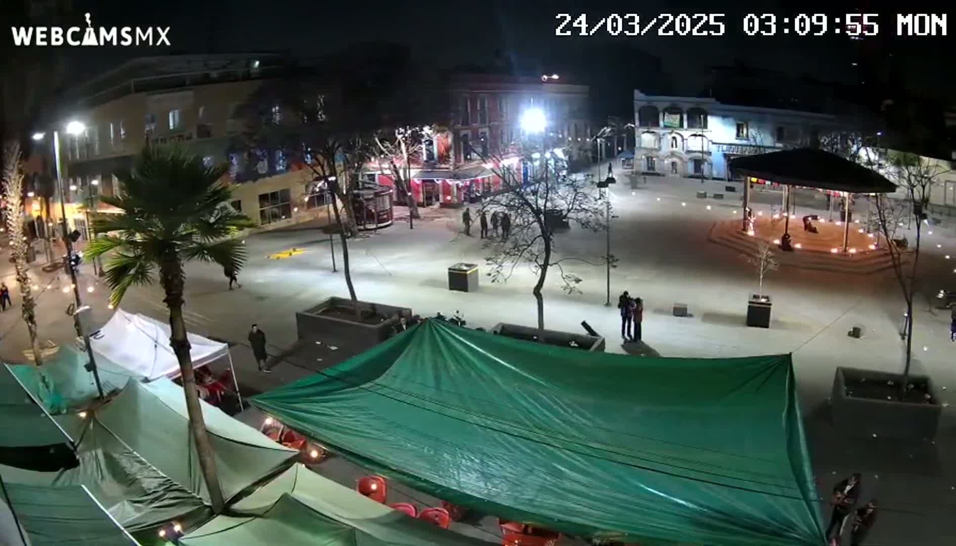 A nighttime view of a town square with several people, some temporary market stalls, and a gazebo in the center, under clear skies.