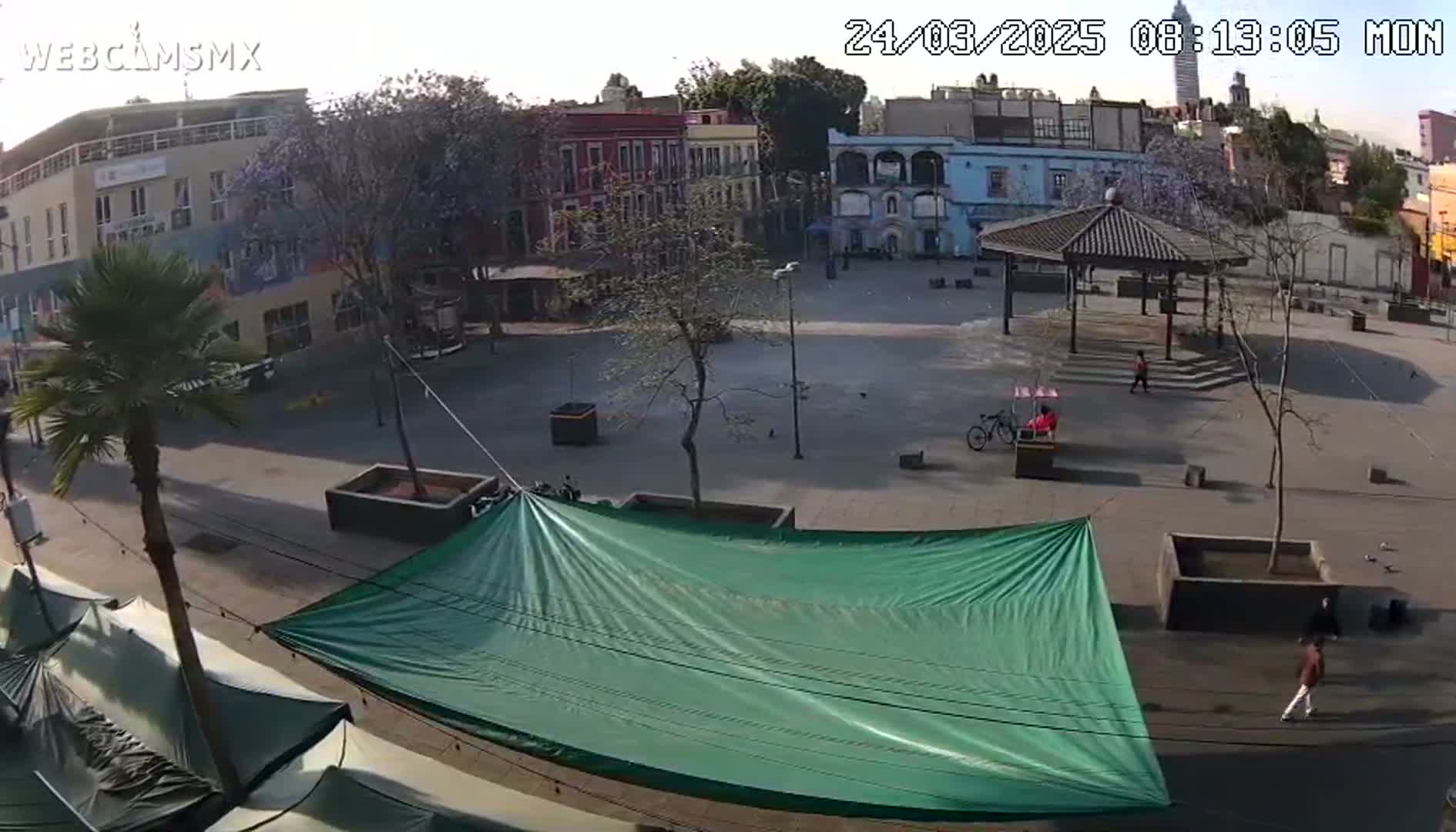 A mostly empty plaza, under a mostly sunny sky, is viewed from above, showing several low buildings, a gazebo, a large green tarp, and a few pedestrians.