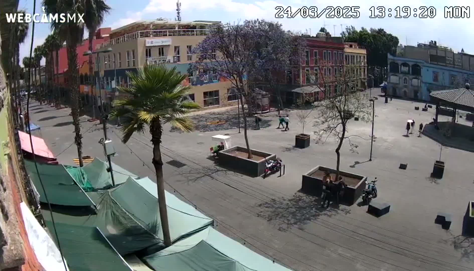 A sunny day in a town square shows several buildings, palm trees, a few people, and some temporary market stalls.