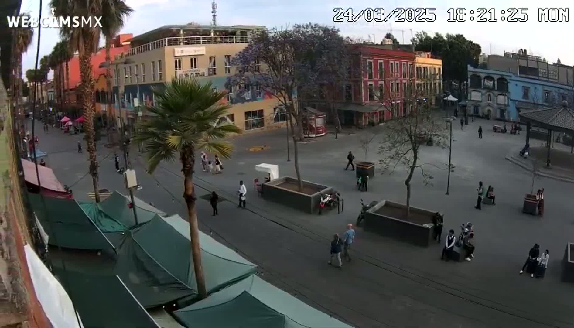 A mostly empty town square with several buildings, palm trees, and market stalls under a partly cloudy sky.