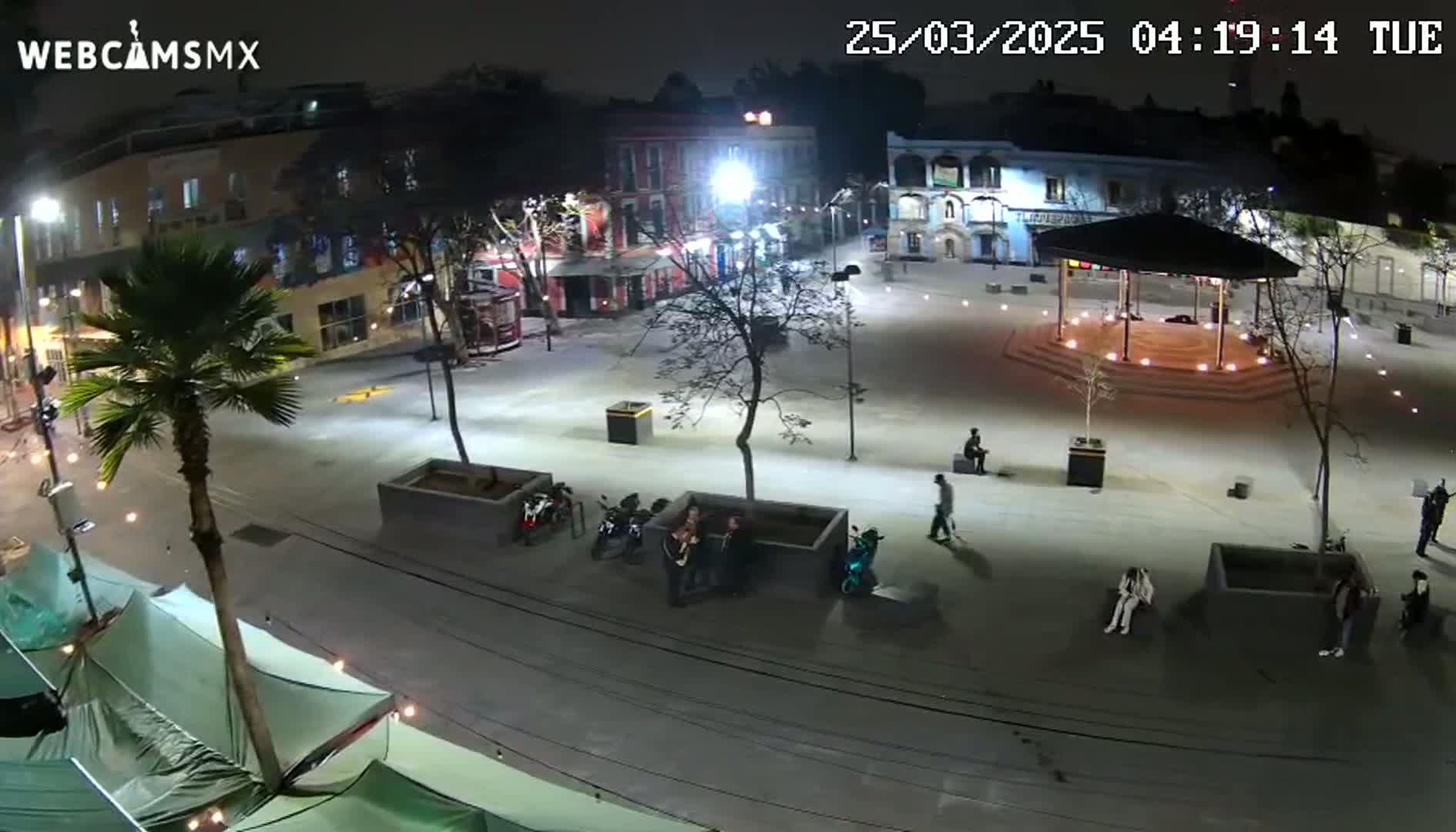A nighttime aerial view of a town square with several people, some parked motorcycles, and a gazebo under bright artificial lights.