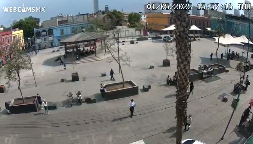 A mostly empty plaza with several people walking around, some sitting in groups, under a sunny sky.