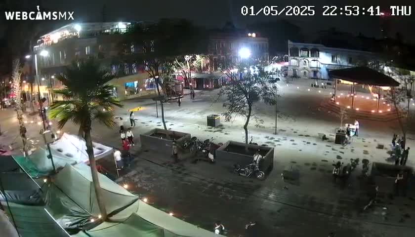 A nighttime, outdoor scene shows a town square with several people, parked motorcycles, and tents under palm trees, with a gazebo in the center.