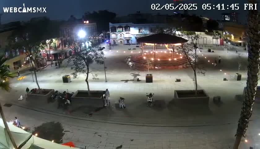 An empty plaza at night, illuminated by lights, with a few people and some parked motorcycles, under a clear sky.