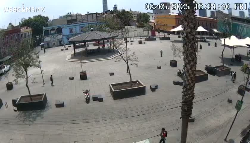 A mostly empty plaza with buildings and trees surrounding it on a sunny day, featuring a gazebo, planters, and a few people.