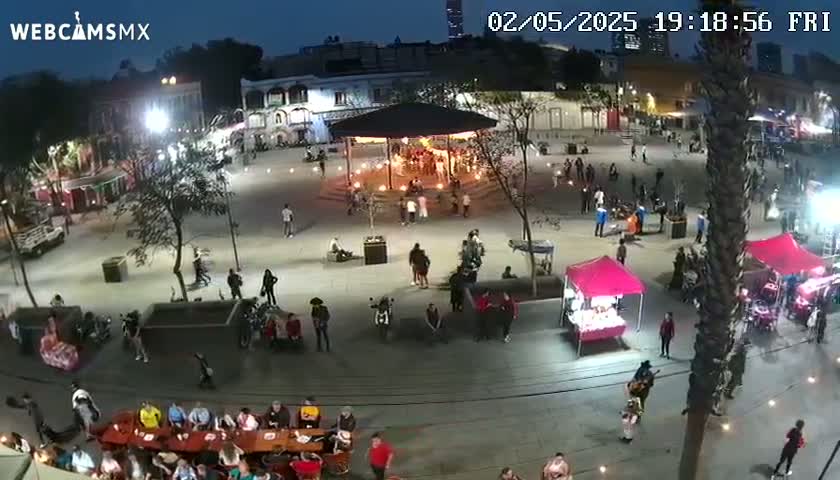 A nighttime aerial view of a town square with people gathered around food stalls and a gazebo, under clear skies.