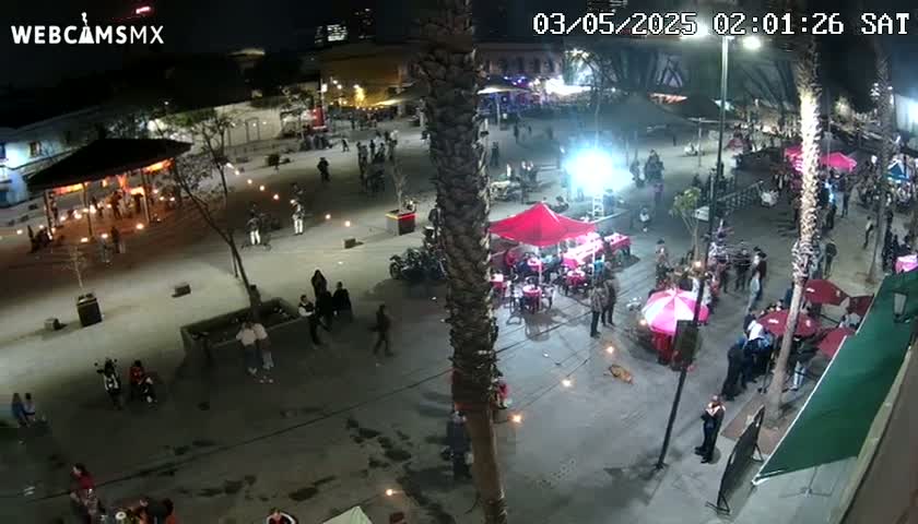 A nighttime aerial view shows a plaza with numerous people gathered around food stalls and tables, under partly cloudy skies.