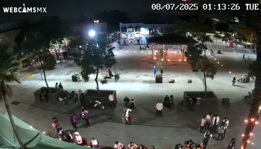 A nighttime, brightly lit plaza with several people gathered in groups and walking around, near buildings and trees.