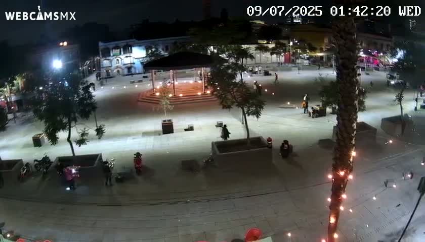 A nighttime, brightly lit plaza with a gazebo in the center, surrounded by buildings, trees, and a few people, some on scooters.