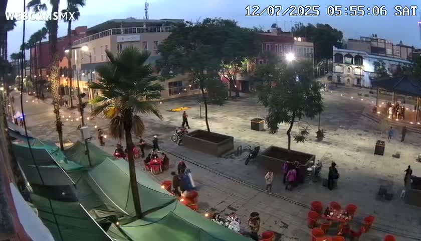 A nighttime, brightly lit plaza with a gazebo in the center, surrounded by buildings, trees, and a few people, some on scooters.
