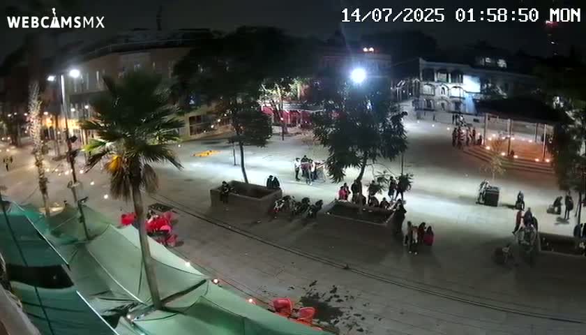 A nighttime, brightly lit plaza shows groups of people gathered in various locations, with palm trees and buildings surrounding the paved area.