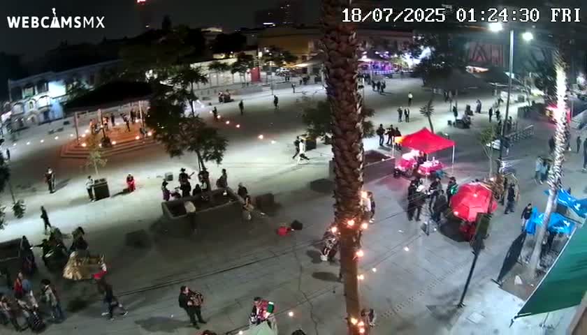A nighttime, aerial view of a plaza with people gathered in groups, some performing music, under lights with a tall palm tree in the foreground.