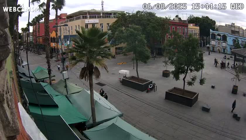 A mostly empty plaza with a few people and several green tents is viewed from above on an overcast day, surrounded by colorful buildings and palm trees.