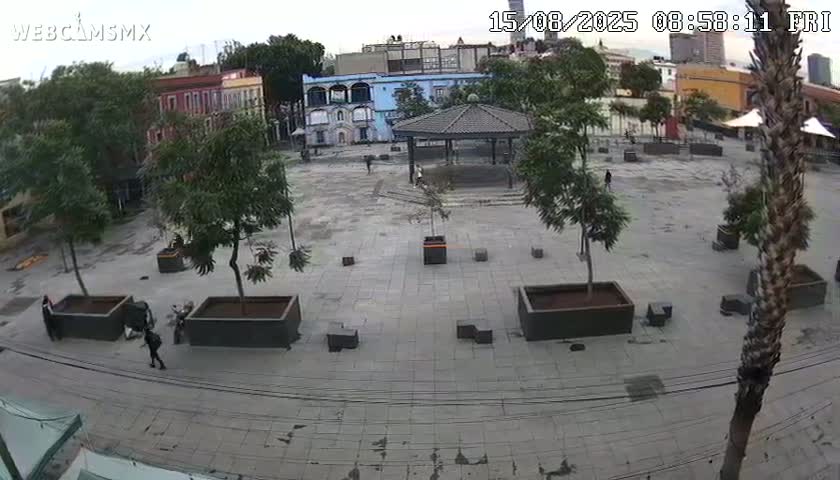 A mostly empty plaza with a few people and several green tents is viewed from above on an overcast day, surrounded by colorful buildings and palm trees.