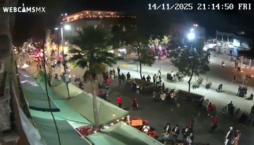 An elevated nighttime view shows a lively outdoor plaza or street, bustling with pedestrians among green awnings and trees, brightly lit by streetlights under clear weather.