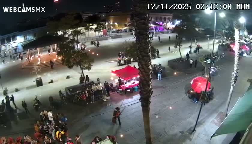 A lively outdoor plaza is depicted at night under clear skies, featuring numerous people, a gazebo, a vendor stall with a red canopy, another large red umbrella, a musician, and bright street lighting.