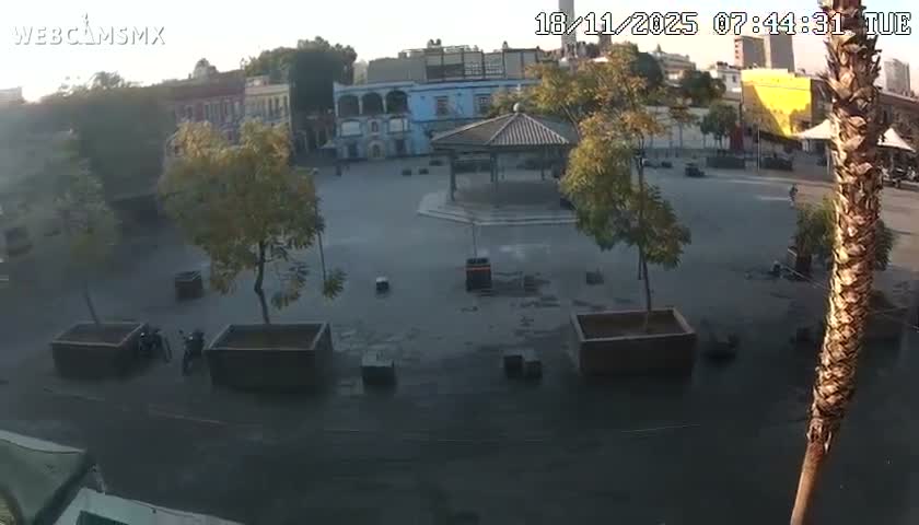 A lively outdoor plaza is depicted at night under clear skies, featuring numerous people, a gazebo, a vendor stall with a red canopy, another large red umbrella, a musician, and bright street lighting.