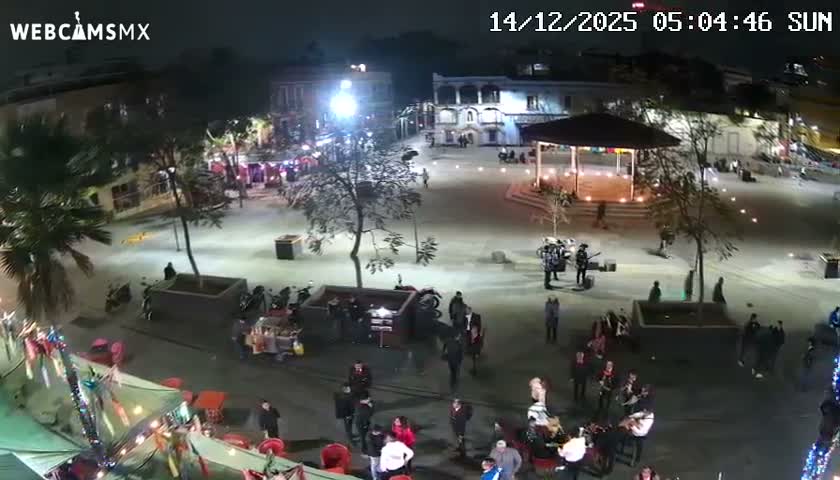 At night, a quiet city street and public square, illuminated by streetlights, feature buildings, palm trees, a gazebo, and a few individuals under a clear sky.