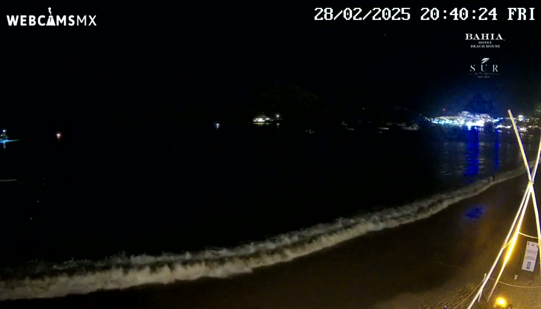 A nighttime beach scene shows gentle waves lapping the shore under a dark sky, with distant lights visible across the water.