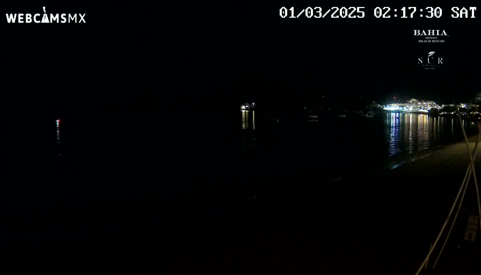 A dark, nighttime beach scene shows a calm body of water reflecting the lights of a town across the water.