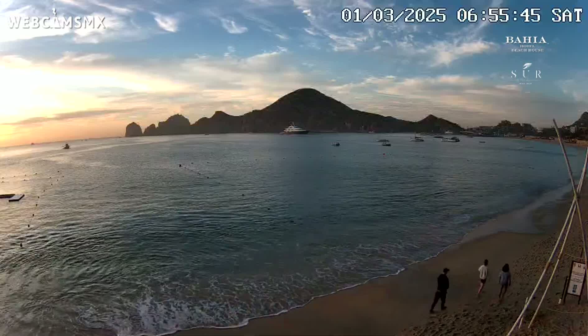 A calm ocean beach at sunset with a few people walking along the shore, and a large yacht in the water near some rocky hills.