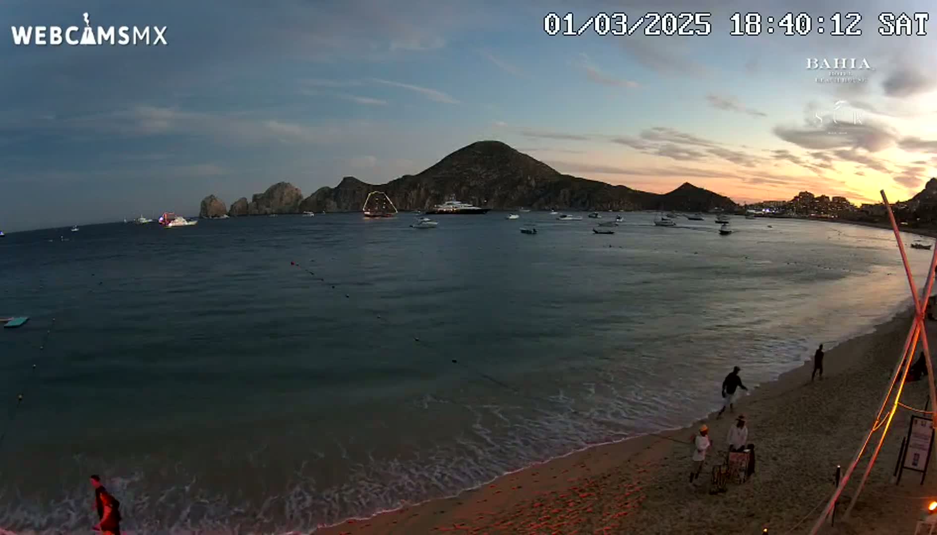 A sandy beach at sunset with calm ocean waves, several boats on the water, and a few people on the shore near some hills.