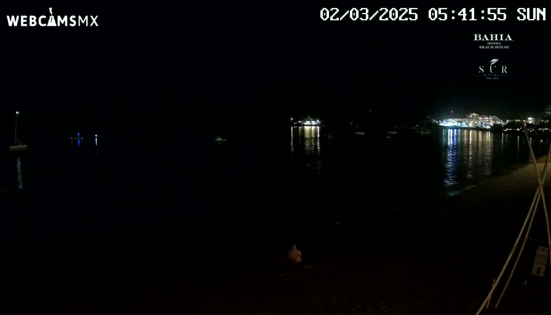 A dark, nighttime beach scene shows a person sitting on the sand near calm water reflecting lights from a distant town.