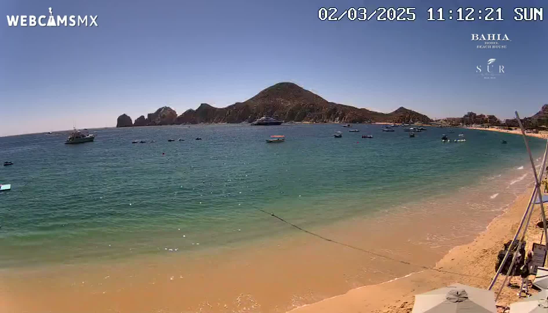 A sunny day at a sandy beach with calm, clear turquoise water, boats, and a mountainous backdrop.