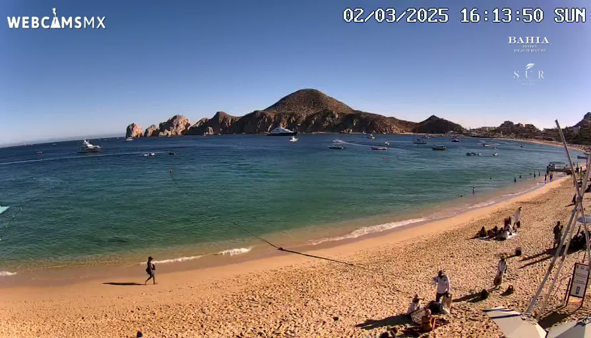 A sandy beach with several people on it and calm, clear turquoise water extends to a bay with boats and rocky hills under a sunny sky.