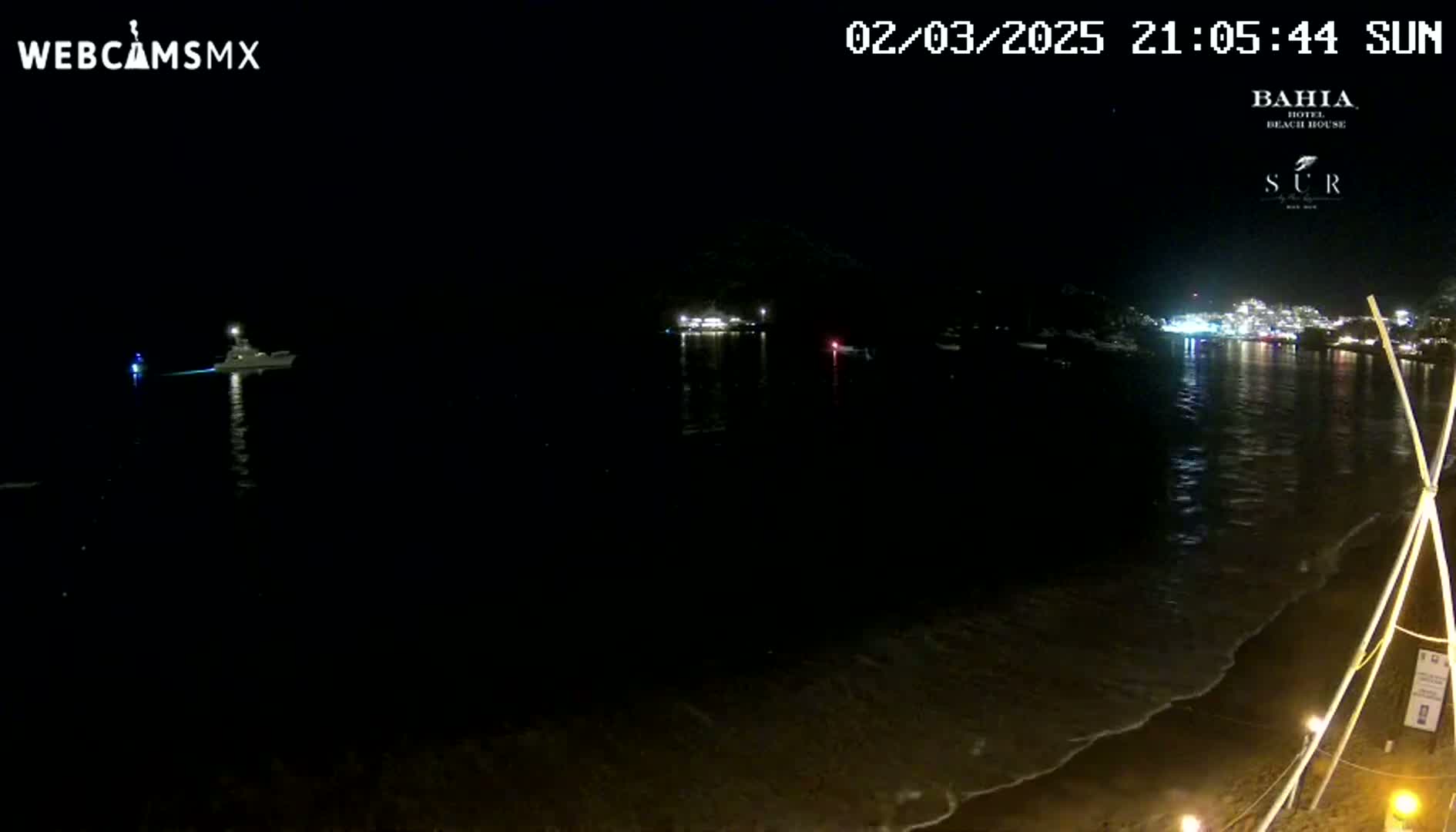 A nighttime view of a dark ocean with a boat, a shoreline with lights in the distance, and gentle waves lapping on the beach.