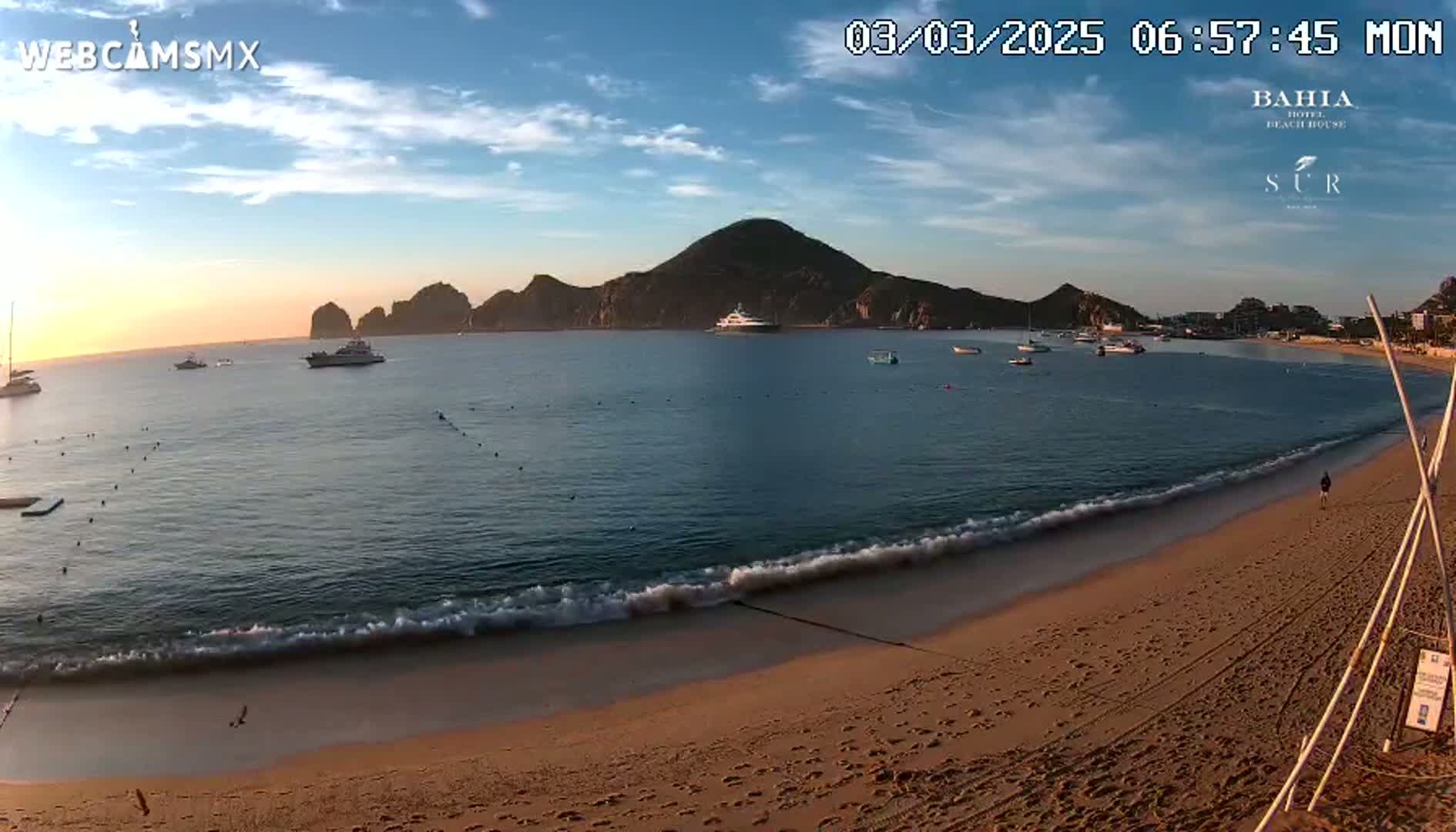 A sandy beach with gentle waves laps the shore under a partly cloudy sunrise sky, with several boats visible in the calm water and a large landmass in the background.