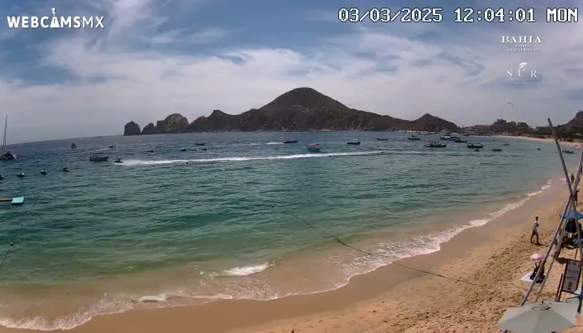 A sandy beach with calm, clear water, numerous boats, and a mountainous backdrop under a partly cloudy sky.