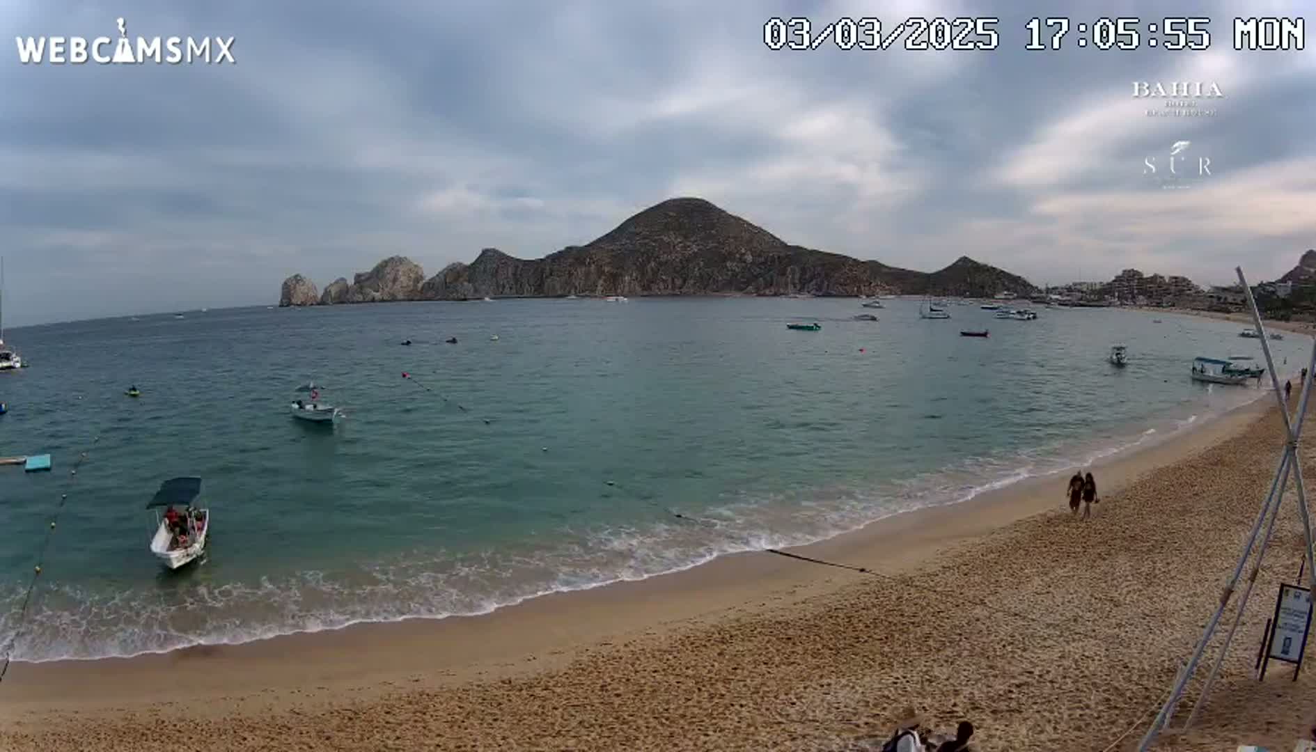 A sandy beach with calm, clear water laps gently at the shore, boats are visible in the bay, and a large landmass rises in the background under a cloudy sky.