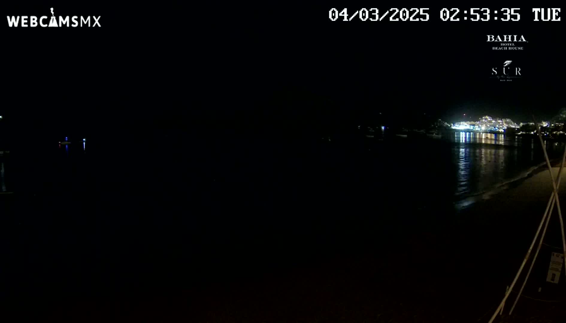 A nighttime view of a dark beach and calm water reflecting the lights of a town across the bay.