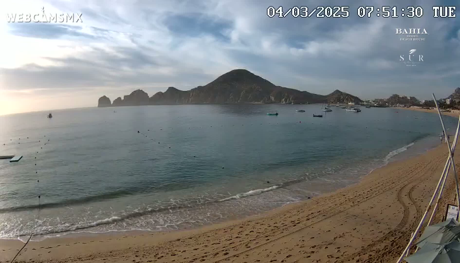 A sandy beach with calm, clear water laps gently at the shore, under a partly cloudy sky, with a large mountain in the background.