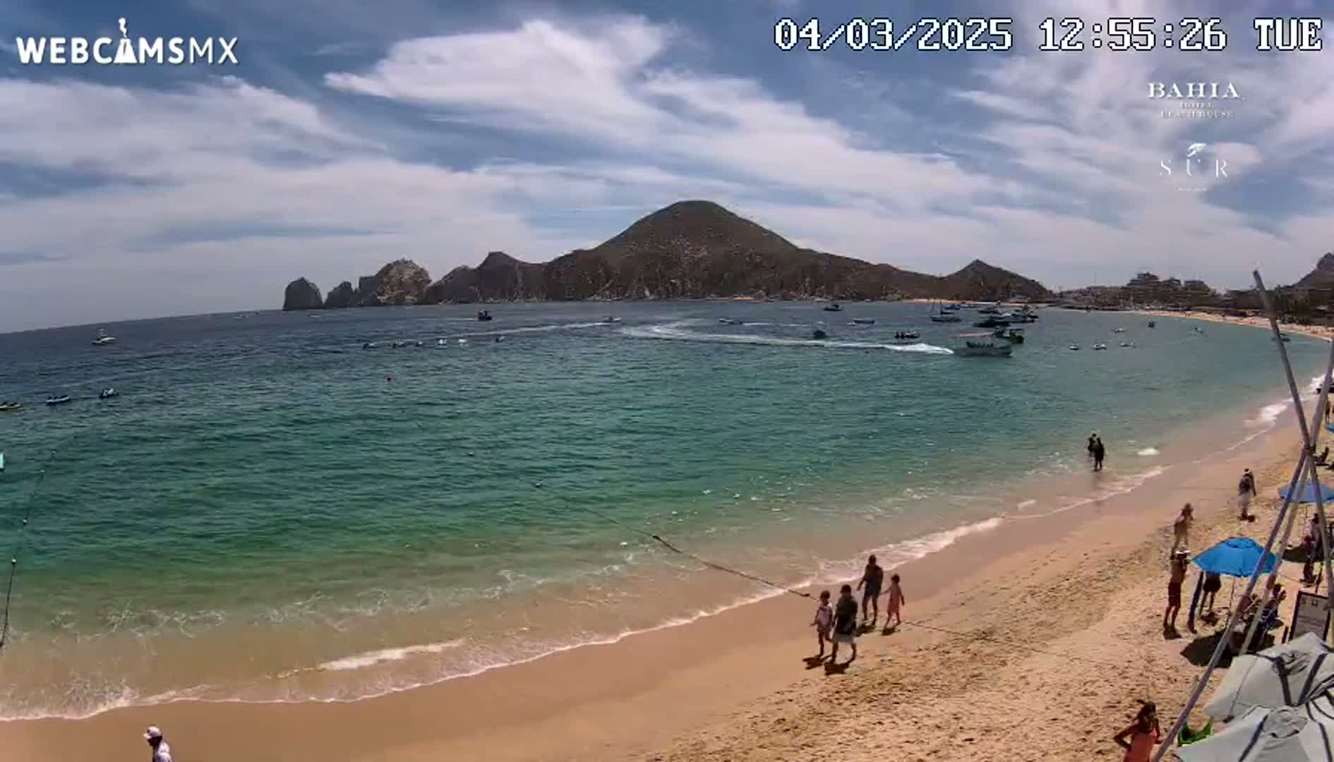 A sandy beach with turquoise water and several people walking along the shore, boats in the bay, and a large rocky hill in the distance under a partly cloudy sky.