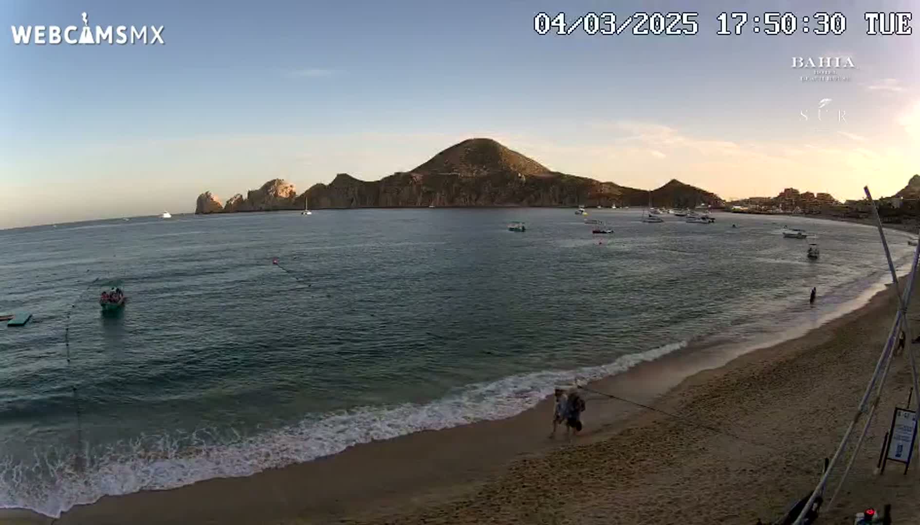 A sandy beach with calm waves laps the shore under a clear sunset sky, overlooking a calm bay with several boats and a mountainous peninsula in the distance.