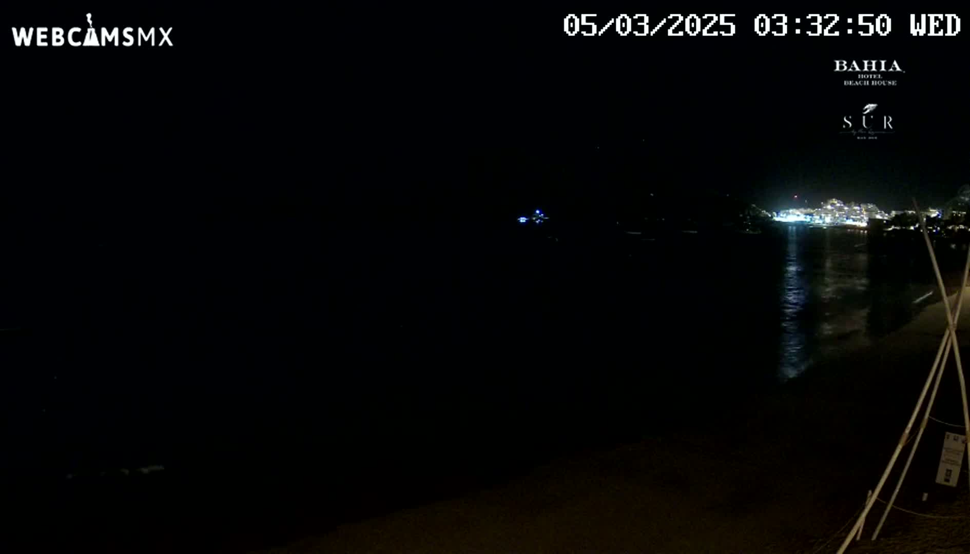 A dark nighttime beach scene shows a calm body of water reflecting the lights of a distant town under a clear sky.