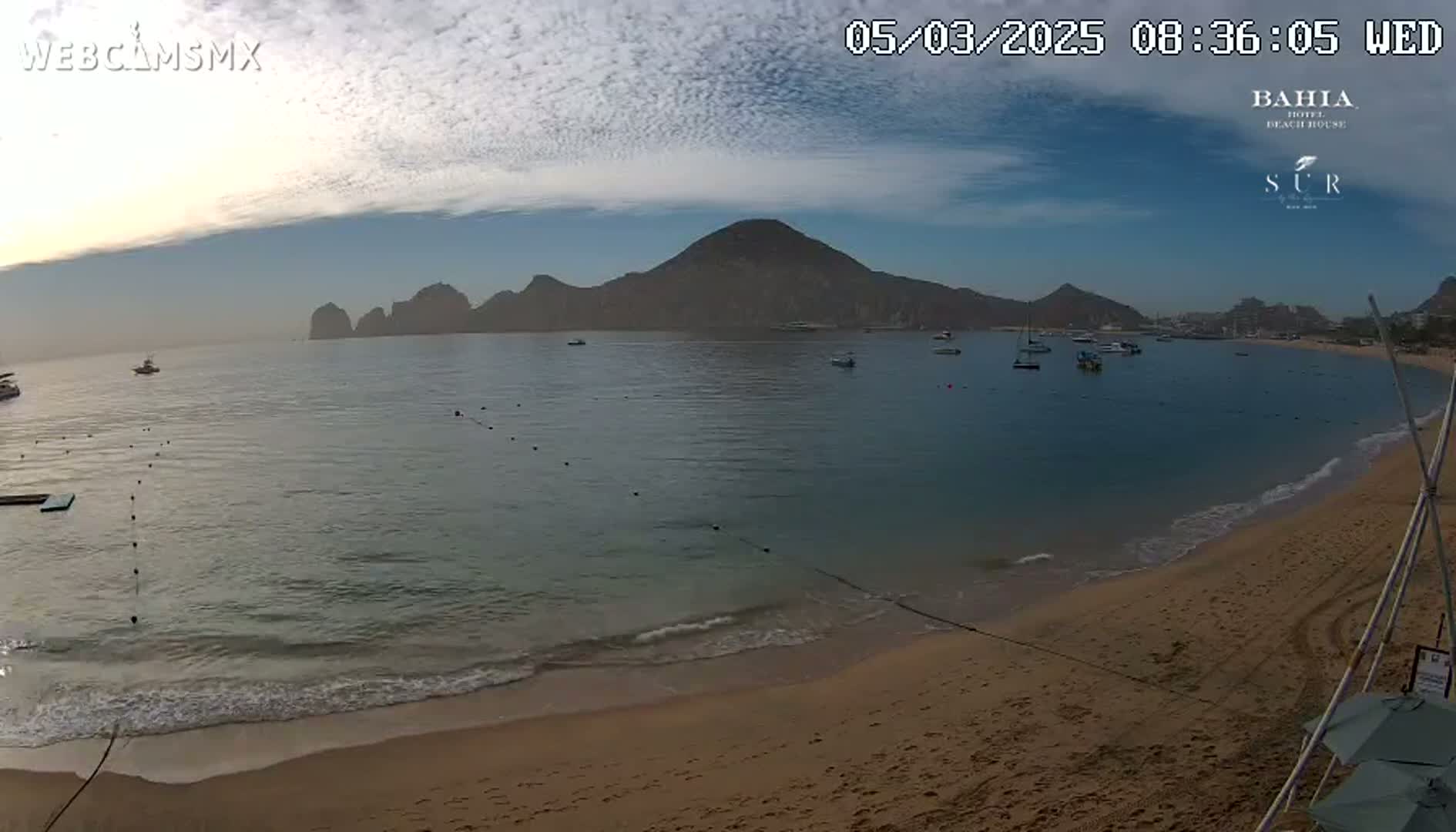 A sandy beach with calm, clear water laps gently at the shore, under a partly cloudy sky, with mountains and boats visible in the distance.