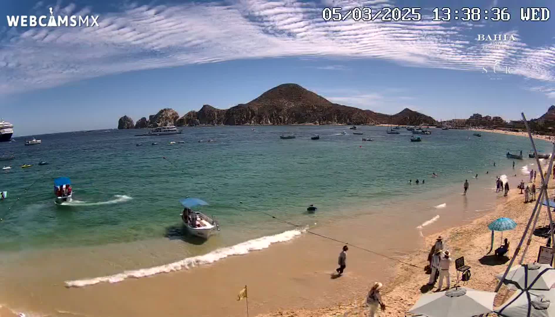 A sandy beach with clear, turquoise water, several small boats, and a large cruise ship in the distance, under a partly cloudy, sunny sky.