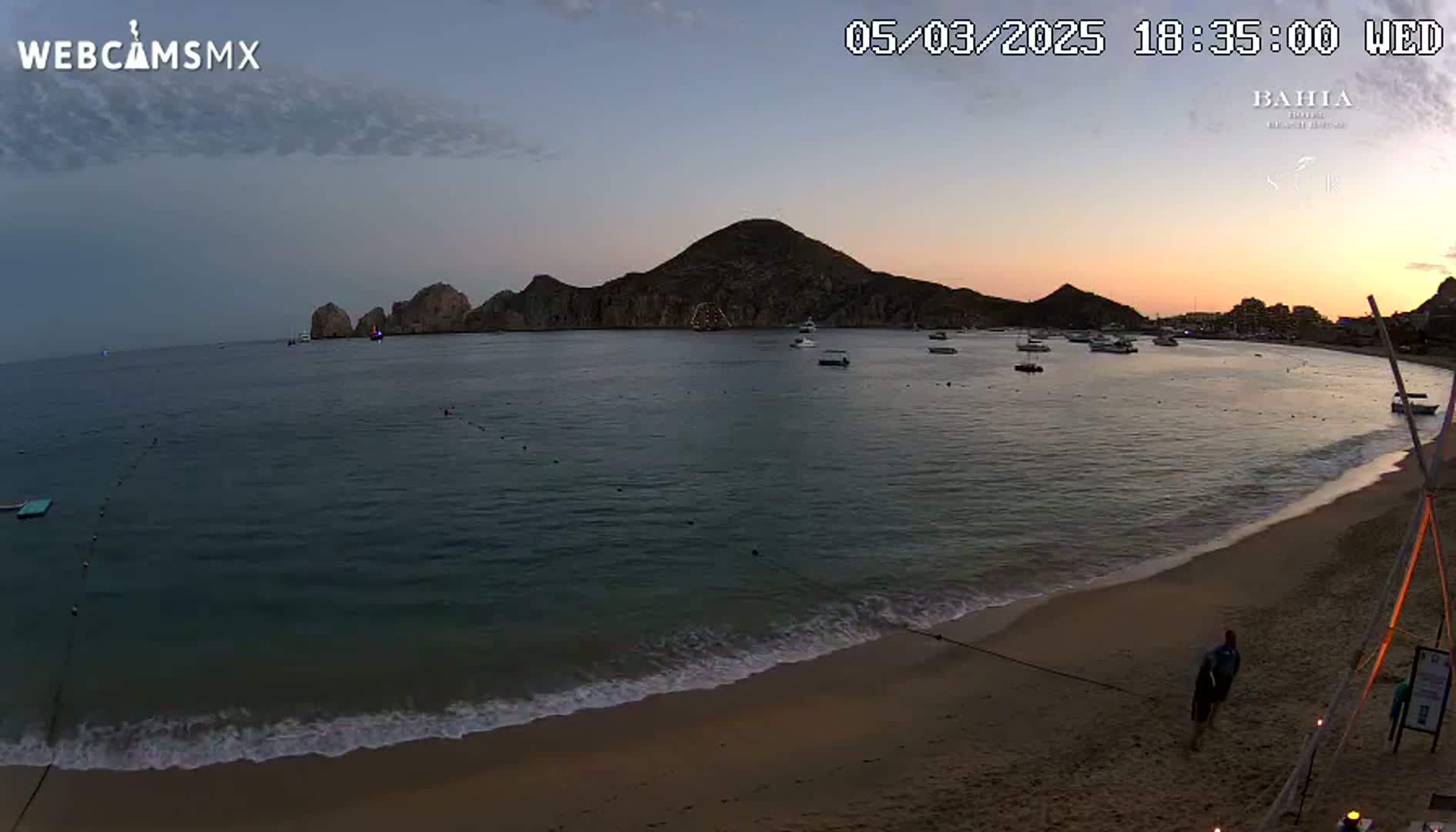 A sandy beach meets calm ocean water under a twilight sky, with a few boats visible in the bay and a rocky hill in the distance.