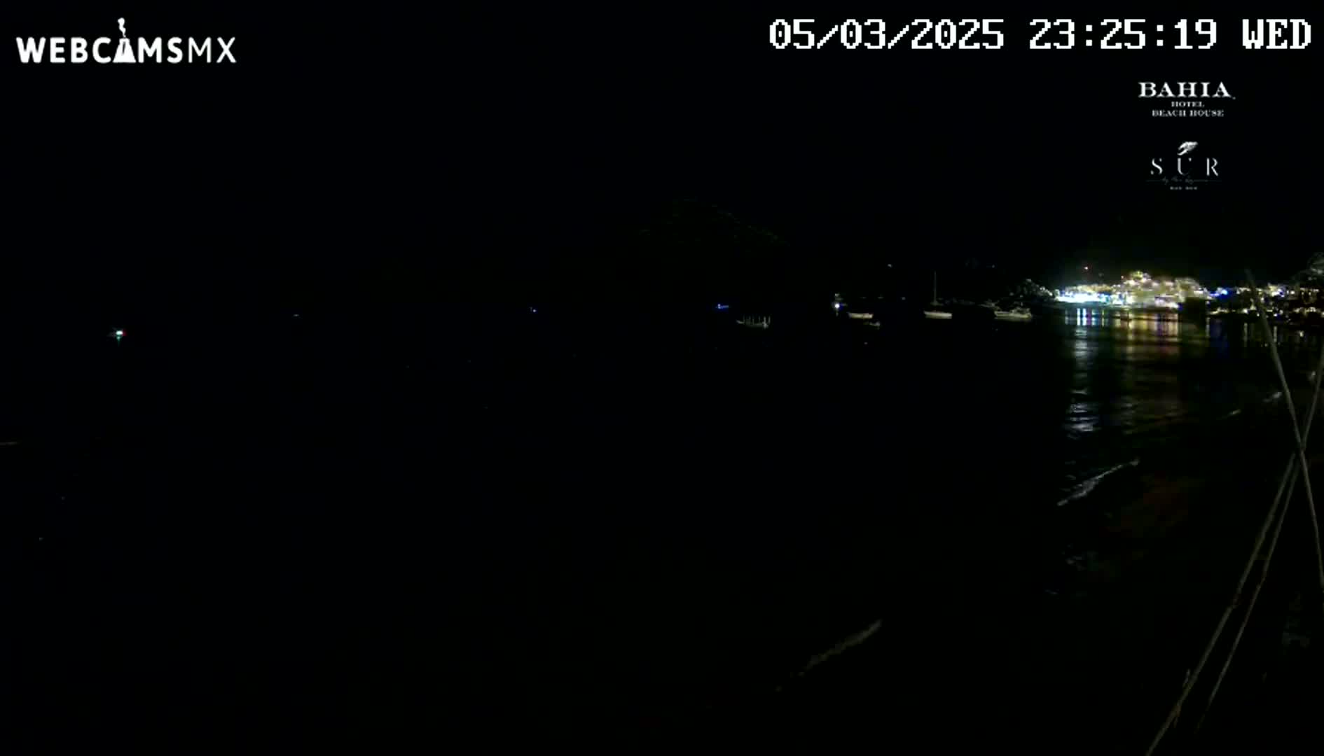A nighttime, dark view of a calm body of water with dimly lit boats and a brightly lit town in the distance.