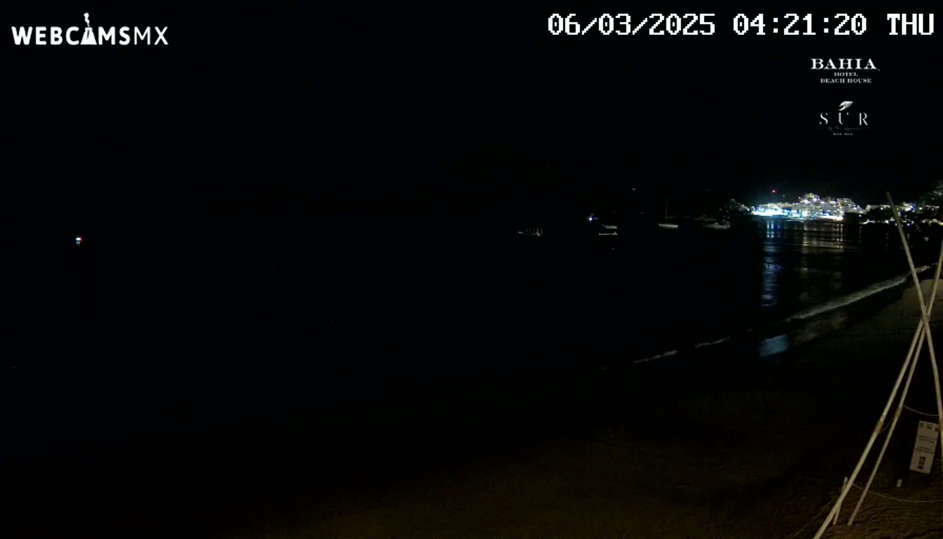 A nighttime beach scene shows dark water reflecting lights from buildings across the bay under a clear, dark sky.
