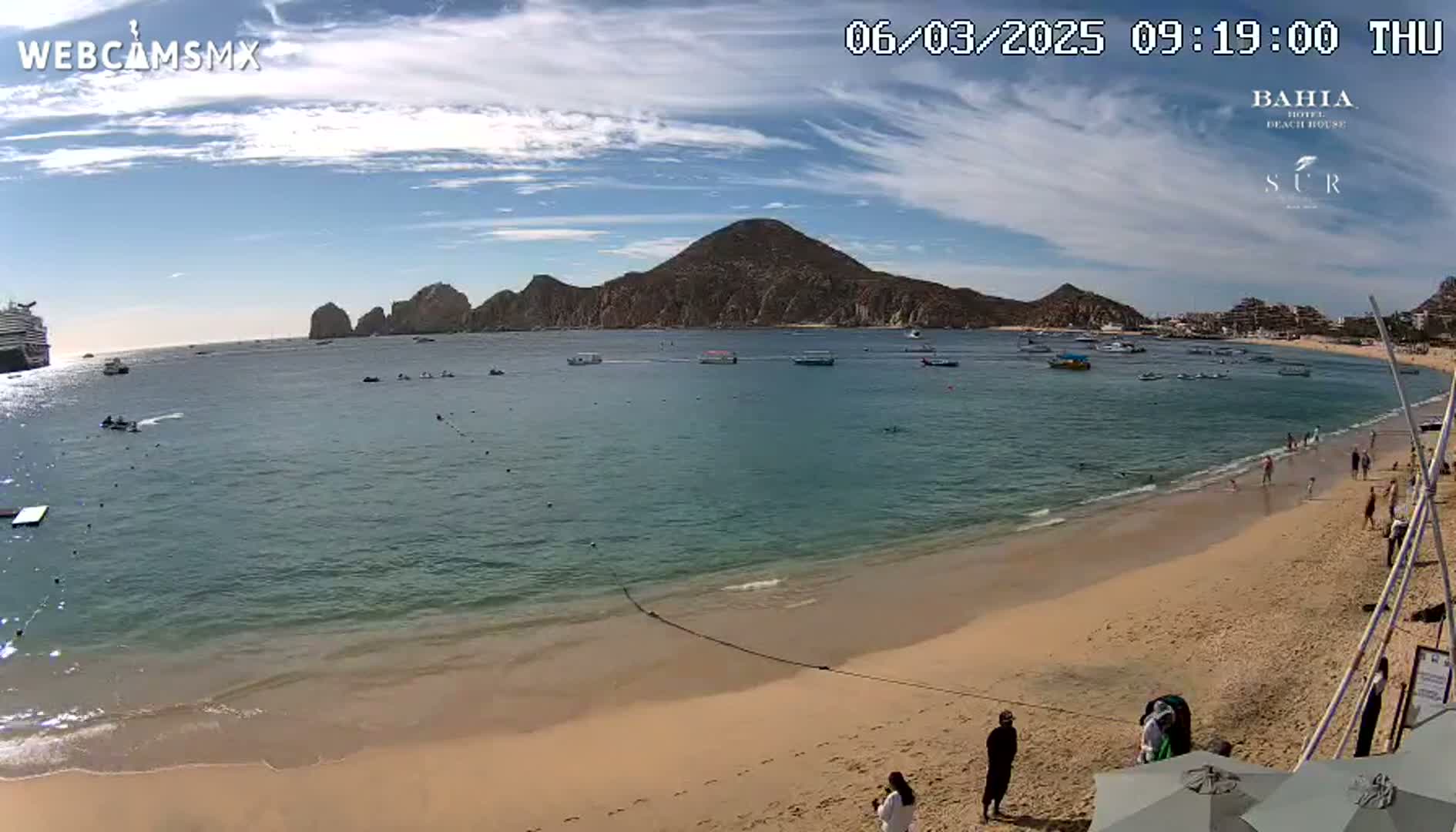 A sandy beach with calm, clear water, boats in the bay, and a large cruise ship in the distance, under a mostly sunny sky.