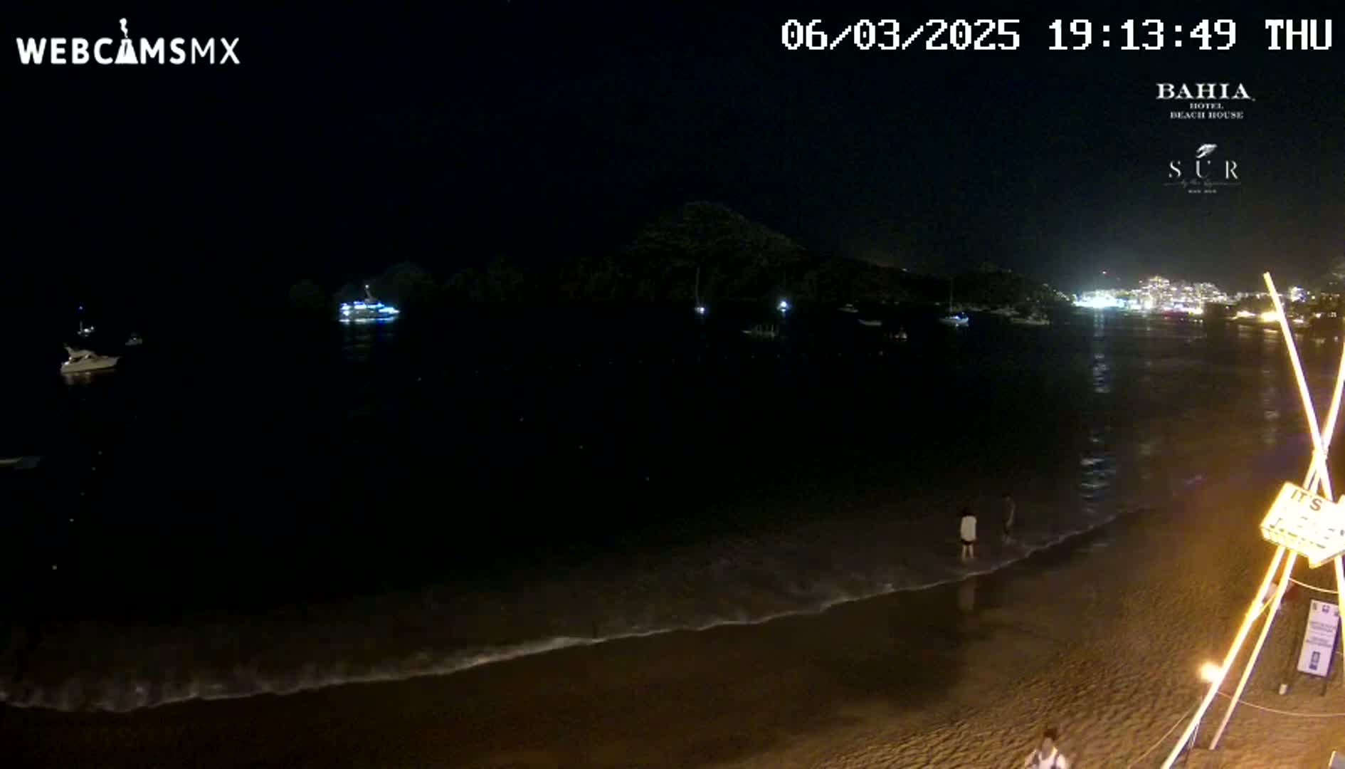 A dark nighttime beach scene shows a calm ocean with gentle waves lapping at the shore, a few boats in the distance, and some people on the beach near a lighted structure.